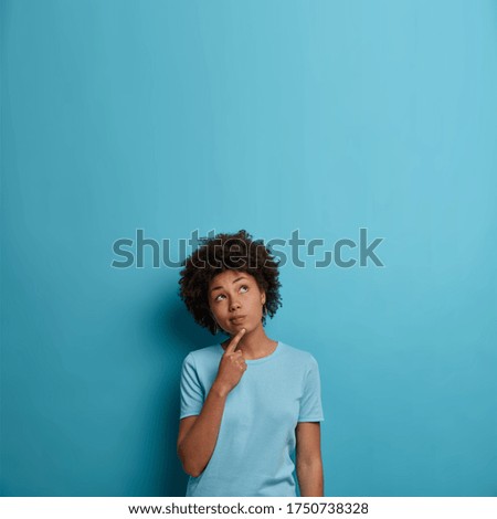 Photo of pensive dark skinned woman keeps finger on chin, concentrated above and ponders about something, wears t shirt, poses against blue background, empty space for your promotion or information