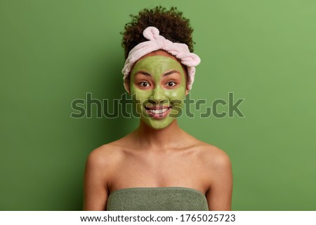 Complexion care and beauty concept. Positive woman with combed curly hair, headband, applies green moisturizing mask on face, has morning skin care routine after taking shower, isolated on green wall