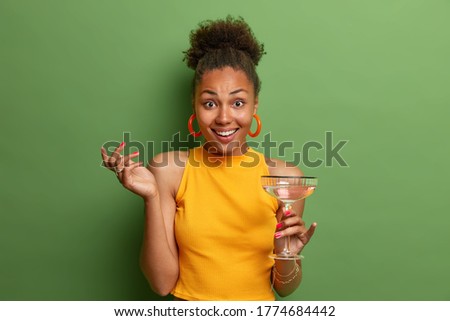 Positive good looking ethnic female poses with glass of summer cocktail, has happy look and nice talk with friend, wears yellow clothes isolated on green background. People, drinking, leisure concept