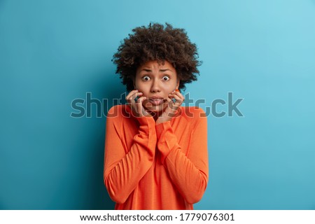 Photo of nervous scared woman grabs face and looks with worried expression at camera, sees phobia, afraids of speaking, wears orange jumper, isolated on blue background. Human reaction concept