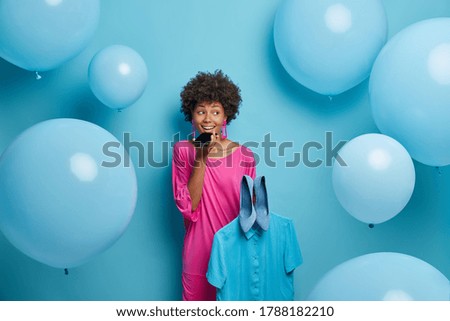 Positive talkative woman makes voice call, consults with friend what better to wear for theme party, holds blue shirt and shoes, dressed in pink dress, poses indoor against big helium balloons