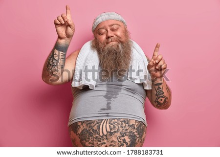 Obesity and sport concept. Joyful overweight man dances carefree has sweaty body tattooed arms points upwards isolated on pink background, does exercises at home, burns calories after eating fast food