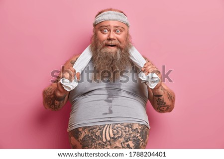 Portrait of fat bearded man with sweaty body, feels tired after exhausting exercises in gym, has big belly sticking out from t shirt, keeps hands on towel, does sport regularly to loose weight
