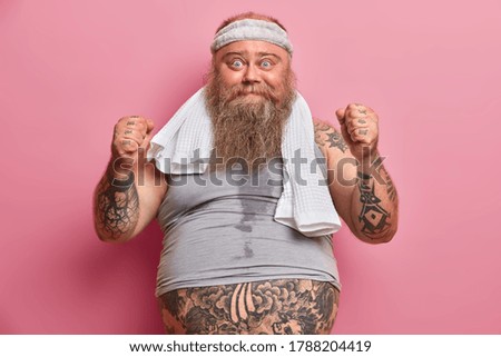 Funny overweight man being sweaty after intensive cardio, raises clenched fists, dressed in sportswear, does morning exercises to loose weight puts all efforts to be fit and healthy. Sport and obesity