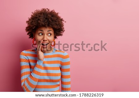 Surprised curious curly woman keeps hand near mouth and whispers secret, spreads rumors, looks with wondered expression aside, dressed casually, isolated on pink background, blank empty space