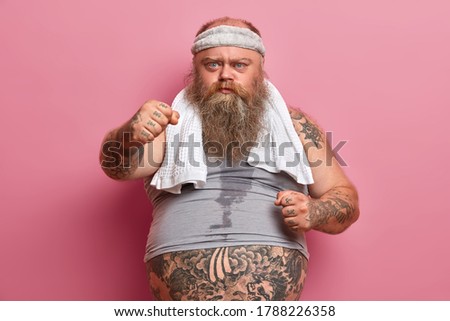 Serious chubby bearded man shows clenched fists, suffers from extra weight, goes in for sport, has sweaty body and tattooed arms, poses against pink background. Slimming and dieting concept.