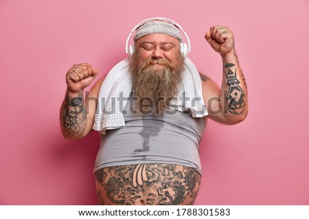 Happy male fatso enjoys workout with music, raises hands and dances, has sweaty body, wears shirt and towel around neck, isolated on pink background. Chubby athlete happy to achieve great results