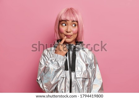 Asian woman has dyed pink hair, looks curiously aside, smiles, wears stylish silver coat, rosy background