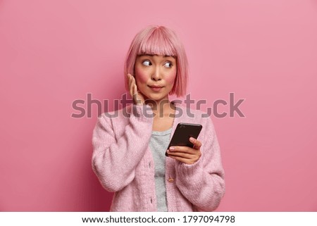 Young Asian woman with surprised face expression looks aside, dressed in casual wear, holds cellphone, surfes social media, sends content shares multimedia online, isolated over rosy background