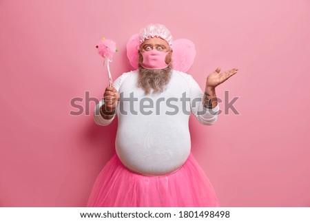Online upbringing during quarantine. Shocked man wears fairy clothes, pink mask, holds magic wand, tries to entertain child via internet, stands indoor against rosy wall, stays at home, feels hesitant