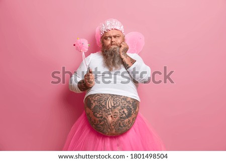 Serious male tooth fairy with big tattooed belly, wears rosy wings, holds magic wand and looks thoughtfully aside, thinks how to entertain childen during holiday, isolated on pink background