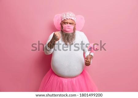 Strict angry fairy man clenches fist, demands staying at home during quarantine time, wears protective mask, wears butterfly wings, pleated pink skirt, poses with magic wand, enjoys childrens holiday