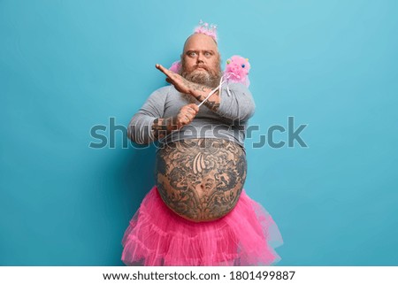 Photo of serious bearded man with big belly, makes refusal gesture, wears fairy costume, refuses to go on stage in funny outfit, crosses hands, poses against blue background. Party time concept