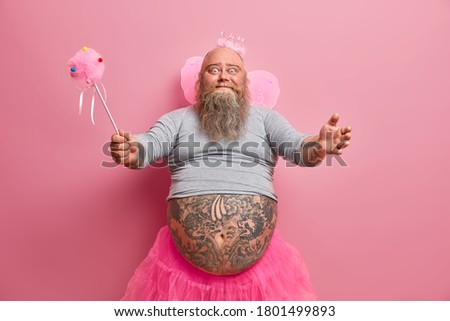 Studio shot of friendly looking kind fatso man wears kids costume for new year party, dressed in fairy outfit, spreads hands and wants to hug someone, isolated on pink background. Happy entertainment