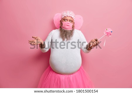 Puzzled hesitant plump father wears masquerade fairy costume, amuses children while staying at home, tries to choose next game, poses with magic wand, rosy background. Holiday, theme party concept