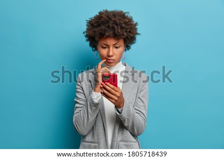 Puzzled confused dark skinned woman in formal wear looks indignant at smartphone, gazes at display, reads business news on website, isolated blue background. People and modern technology concept