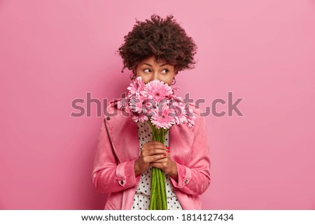 Studio shot of good looking curly haired woman smells flowers, enjoys pleasant odor and stands in stylish clothes indoor. Lovely African American lady holds beautiful bouquet of gerbera daisy