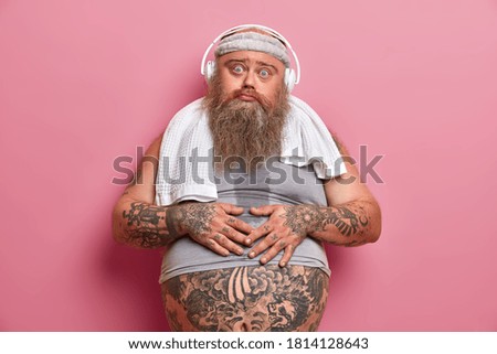 Horizontal shot of displeased overweight man keeps hands on fat belly, feels tired after fitness training and listens music from playlist in headphones. Obese guy promots sport participation