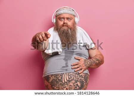 Horizontal shot of angry bearded man points at you with irriatated expression, feels angry after training session in gym. Fatty male wears undersized shirt, listens music and works on losing weight