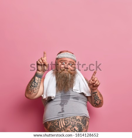 Vertical shot of bearded Caucasian adult man gets sweaty during his training session, works hard on losing excess weight and points above. Funny obese guy wears headband shirt, goes in for sport