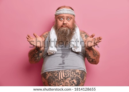Puzzled bearded man with excess weight, spreads palms and looks confused at camera. Fatso guy faces dilemma how to loose weight. Sweaty overweight European adult male has big tattooed belly.