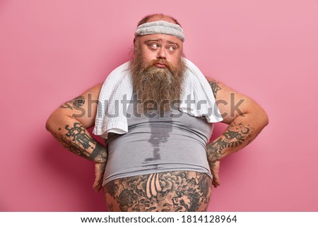Middle age sad man with thick beard keeps hands on hips and looks aside, takes break after exhausting cardio training. Fatso guy burns calories after nourishing dinner, has sweaty tattooed body