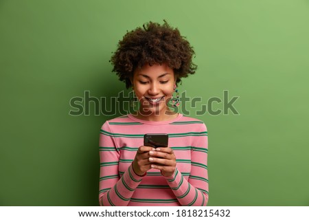 Pretty curly haired woman in striped jumper uses new application on smartphone enjoys high speed internet and types messages. Glad dark skinned teenager uses modern device isolated on green wall
