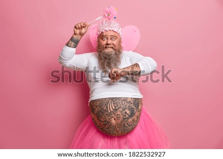 Happy amused bearded European man has fun on carnival or costume party dances with raised arms holds magic wand wears fairy outfit has fat tattooed belly. People holiday celebration concept.