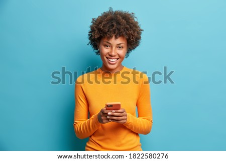 Photo of cheerful dark skinned young woman holds modern device in hands browses her favorite web page surfs social media sends sms smiles positively dressed casually isolated on blue background
