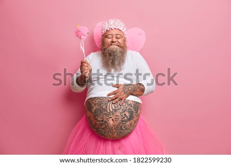 Positive bearded father giggles happily feels satisfaction and plays with kids on holiday holds belly dressed in fairy costume holds magic wand has image of princess isolated over pink background