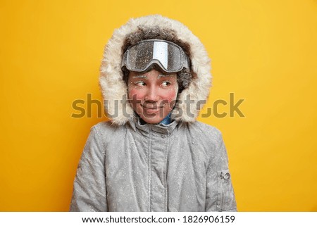 Dreamy winter woman with red frozen face enjoys holiday mountain resort during cold good snow day covered by snowflakes dressed in warm jacket with hood wears ski goggles likes extreme sport.