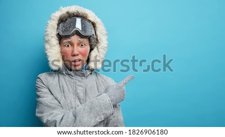 Photo of frozen woman with red frosty face covered by snowflakes wears ski goggles and outerwear indicates at empty space explores polar regions isolated on blue background. Winter destination