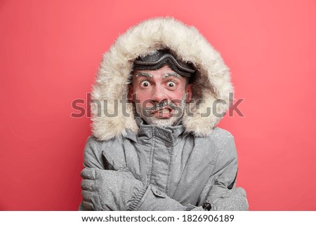 Frozen man trembles from cold has red face covered by ice frosted beard wears jacket with hood needs to warm during winter expedition poses over coral background. Cold weather low temperature