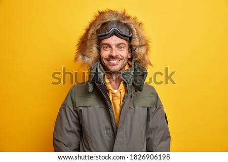 Happy European man in jacket with fur hood feels warm and comfortable during winter time enjoys favorite season smiles happily wears skateboarding goggles has active rest isolated on yellow wall