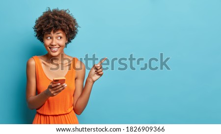Nice attractive young Afro American woman uses cell phone and points on copy space shows direction dressed in stylish orange dress isolated over blue background. Ethnic lady with modern device 商業照片 © 