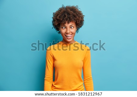 Funny crazy comic Afro American woman makes grimace and crosses eyes plays fool has fun alone sticks out tongue wears casual jumper poses against blue background. Human face expressions concept