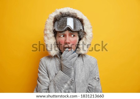 Puzzled ethnic woman with face covered by ice feels cold looks away wears winter jacket gloves freezes during frosty weather isolated on yellow background. Teenage girl goes snowboarding in mountains