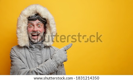 Happy frozen man in warm clothes has fun during winter points away on empty space wears gloves isolated on yellow background. Cheerful male adventurer with frosted beard and ski goggles has expedition