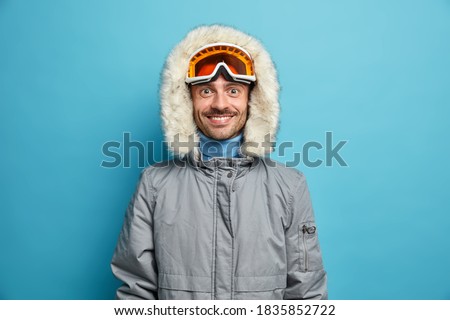 Sporty glad man enjoys winter sport recreation smiles gladfully wears ski goggles and grey jacket isolated on blue. Holidays tour recreation and traveling concept. Snowboarder leads active lifestyle