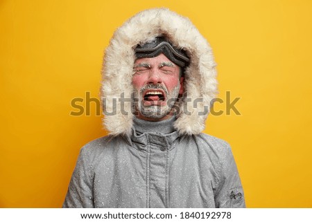 El hombre abandonado llora y se siente infeliz vestido con ropa de invierno se siente muy frío después de que el snowboard tiene la cara roja cubierta de helado con gafas de esquí aisladas sobre la pared amarilla. Concepto de recreación