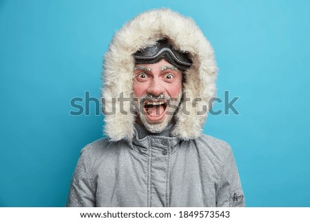 Emotional frozen man shouts loudly has red face covered with ice dressed in thermo jacket with hood and snowboard goggles poses over blue background. Winter guy recreats in mountains during holidays.