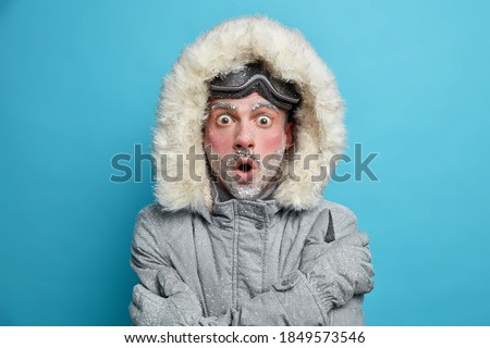 Stupefied frozen man with red face trembles from cold embraces himself to feel warm stares bugged eyes at camera wears snowboarding goggles isolated over blue studio background. Low temperature