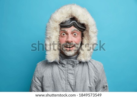 Emotional shocked cheerful man wears winter jacket has red face covered with ice cannot believe in astounding news isolated over blue background. Male snowboarder recreats during cold season