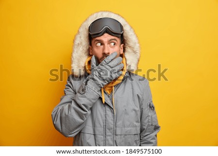 El miedo emocional del hombre snowboard cubre la boca mientras trata de esconder secretos usa guantes cálidos y chaleco de termo gris aislado sobre fondo amarillo aturdido por fuerte tormenta de nieve o baja temperatura