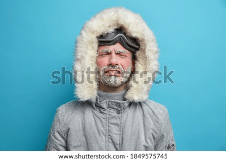 Desperate upset frozen man cries as feels very cold during blizzard and heavy snow storm dressed in thermo grey jacket with hood goes skiing poses against blue background. Dejected snowboarder