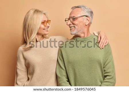 Caring middle aged woman embraces her husband looks with love and broad smile. Married mature couple have good relationships isolated on brown background. Smiling aged husband and wife pose for photo