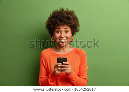 Isolated shot of happy dark skinned woman reads good news via smartphone enjoys mobile application chats online smiles gently uses unlimited internet isolated over green background does shopping