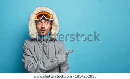 Surprised adult man wears winter jacket and ski goggles has wondered face expression indicates on blank space shows copy space over blue background. Male snowboarder advertises something. Winter time