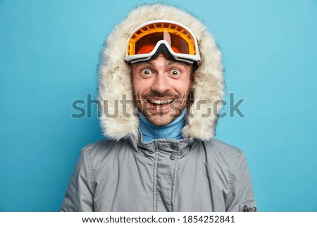 Un hombre alegre y sin afeitar con una expresión expresiva, sonrisas ampliamente, lleva puesto una chaqueta de invierno con gafas de esquí, con capucha, y disfruta de poses de deporte de invierno extremo contra el fondo azul. Concepto de pasatiempo para Snowboard