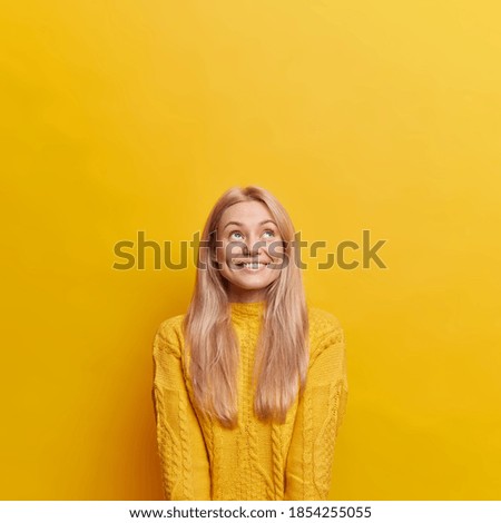 Vertical image of glad young European woman with blonde hair looks above happily has white teeth poses over yellow background empty space upwards for your promotion dressed in casual jumper.